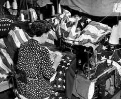 US flags piled up on a table to the left of a female, who is on a sewing machine making a US flag.